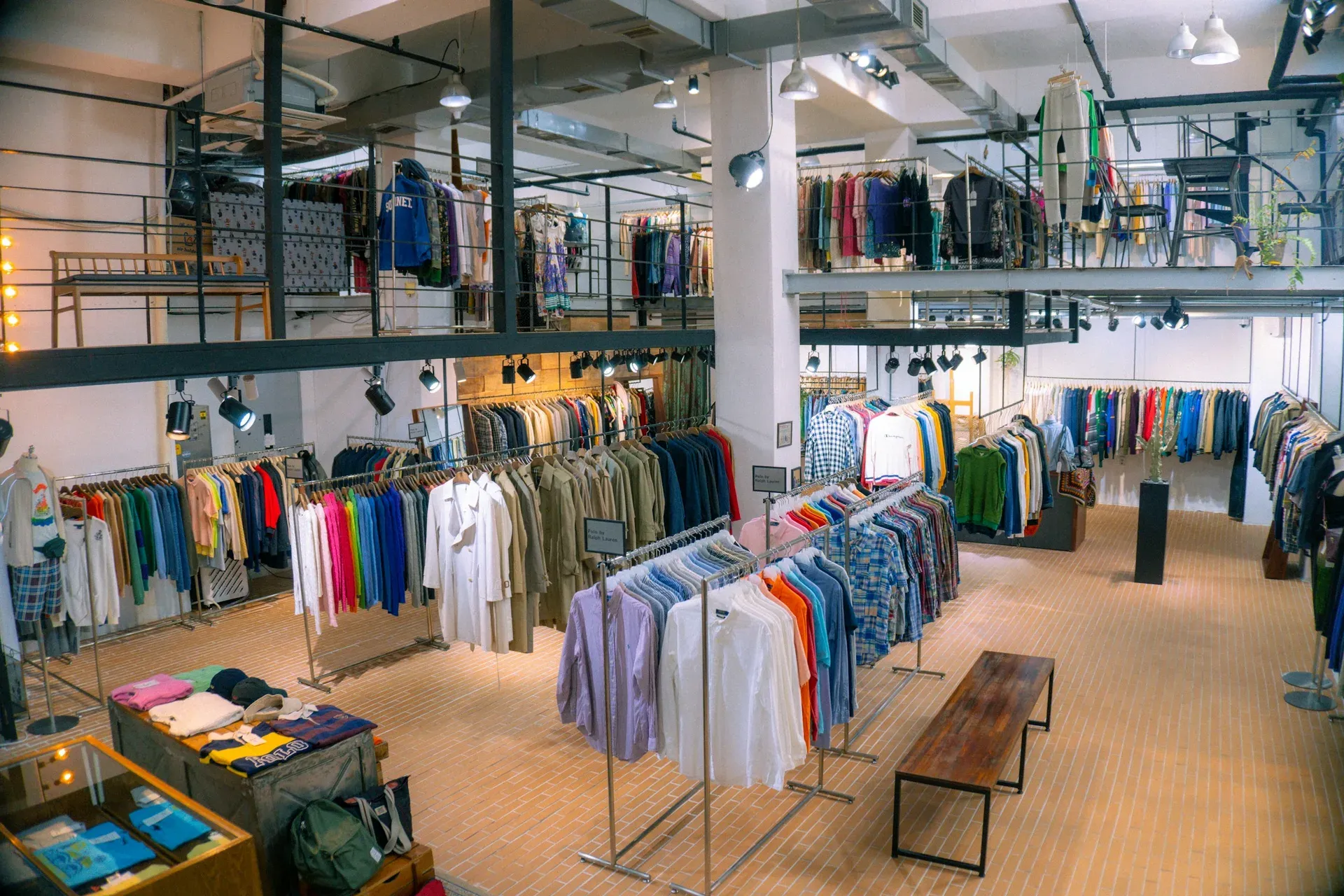 A two-level garment showroom with rails of shirts, knitwear and jackets under industrial pendant lighting